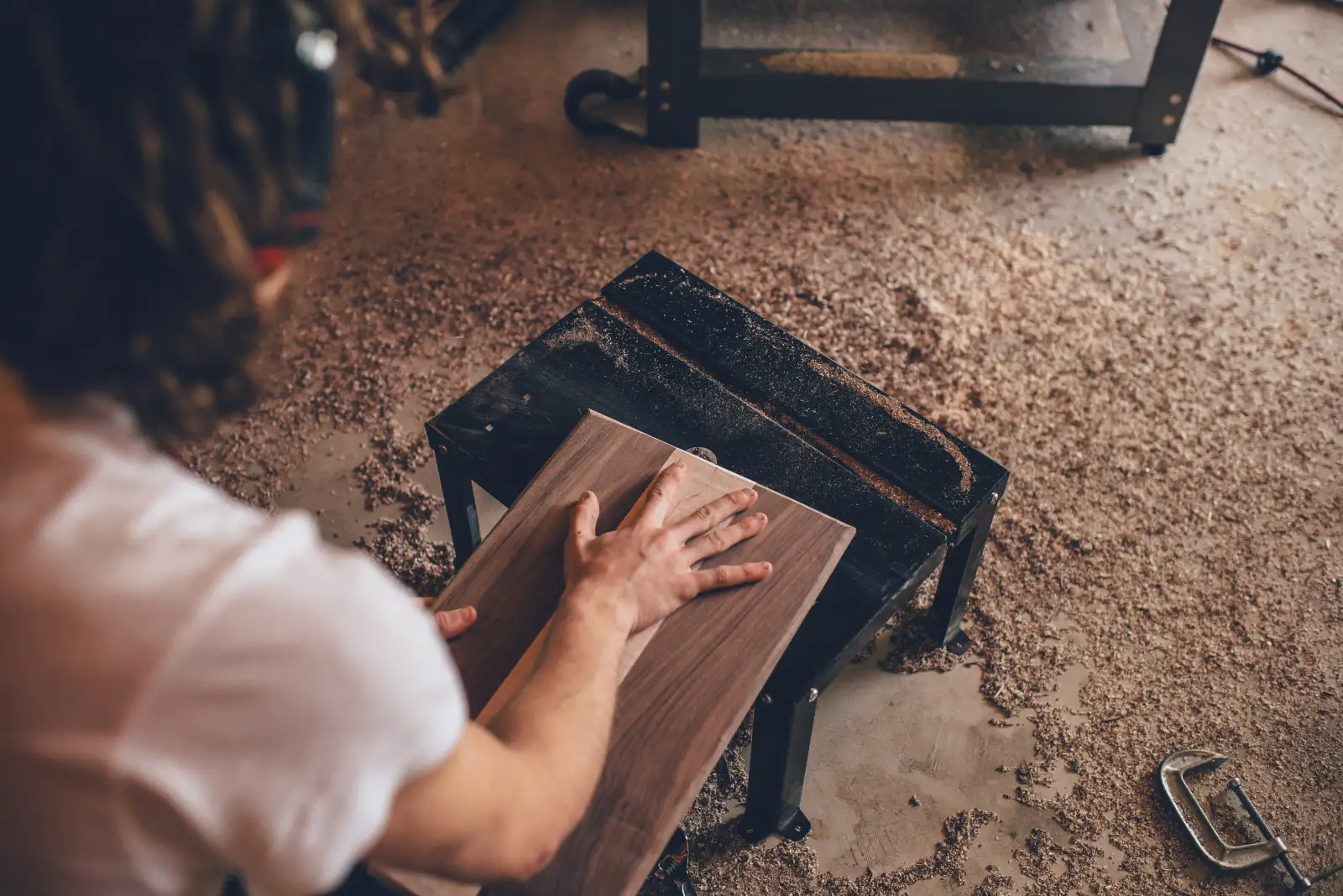 Hands holding a walnut board on a workbench, with sawdust on the floor around it